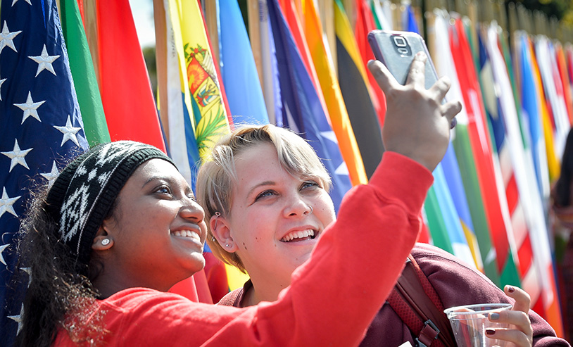 two students taking a photo together with a mobile phone with international flags behind them