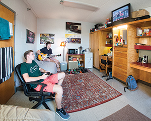 two male students in a dorm, watching tv and playing the guitar