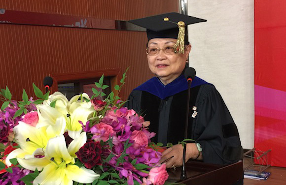 Dr. Lin in graduation regalia, speaking at a Commencement