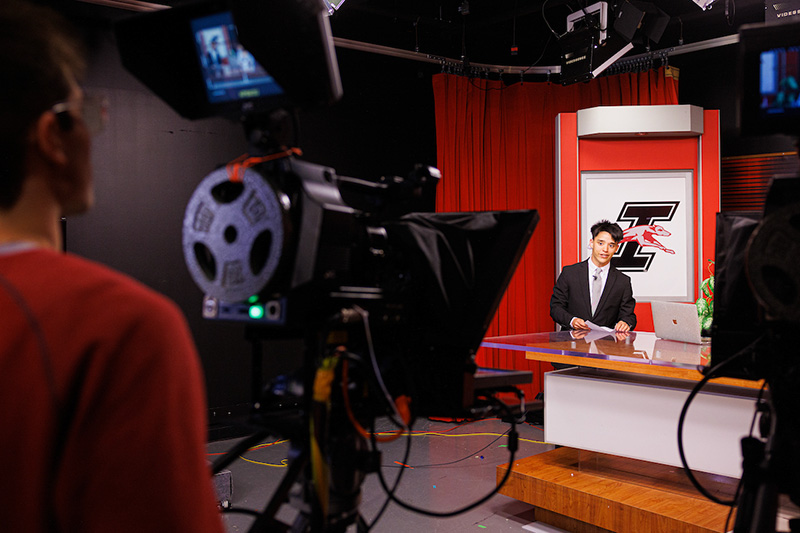 young man sitting behind news desk in front of camera