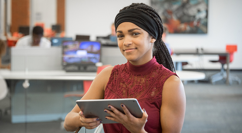 student in library holding tablet smiling