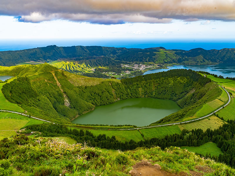 Panoramic view of a green volcanic crater with a lake inside, surrounded by rolling hills and distant coastline under dramatic clouds.