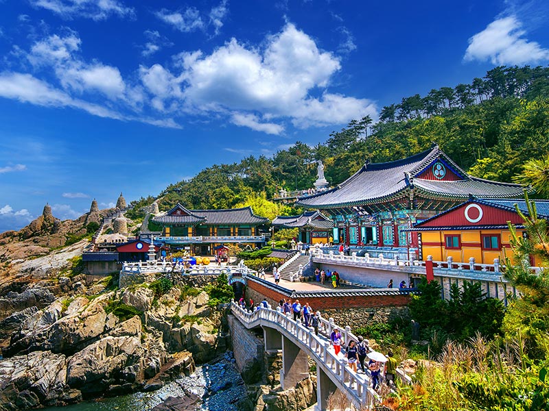 Colorful seaside temple complex on a rocky coastline, with visitors crossing a bridge under a bright blue sky and forested hills.