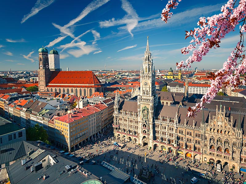Aerial view of Munich’s Marienplatz with the New Town Hall tower and nearby cathedral rooftops under a blue sky, framed by pink cherry blossoms.