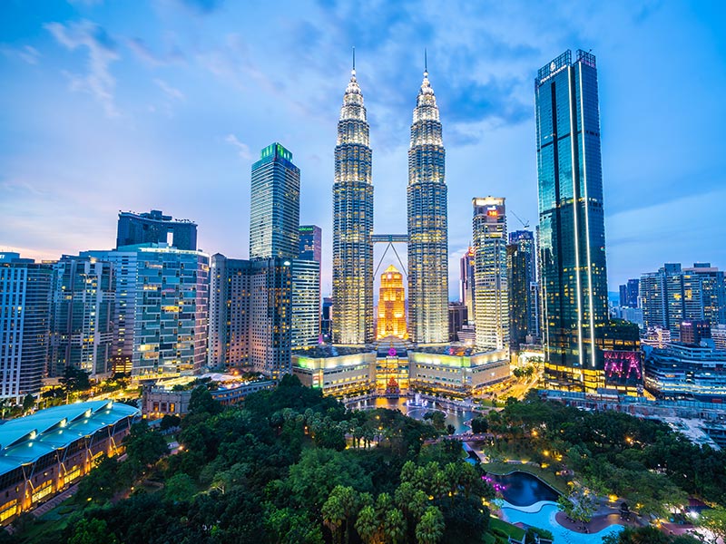 Kuala Lumpur skyline at dusk with the illuminated Petronas Twin Towers centered among modern high-rise buildings and a park in the foreground.