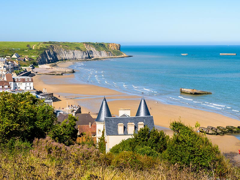 Coastal town overlooking a sandy beach and calm blue sea, with green cliffs curving along the shoreline under a clear sky.