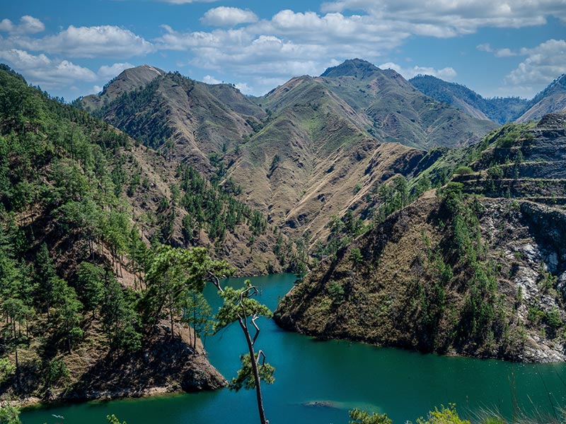 Mountain ridges surround a turquoise lake, with pine-covered slopes under a partly cloudy blue sky.