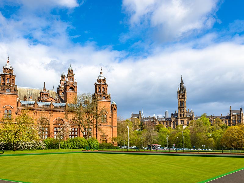 Panoramic view of historic red-sandstone university buildings and a tall Gothic tower beyond a green lawn under a partly cloudy blue sky.