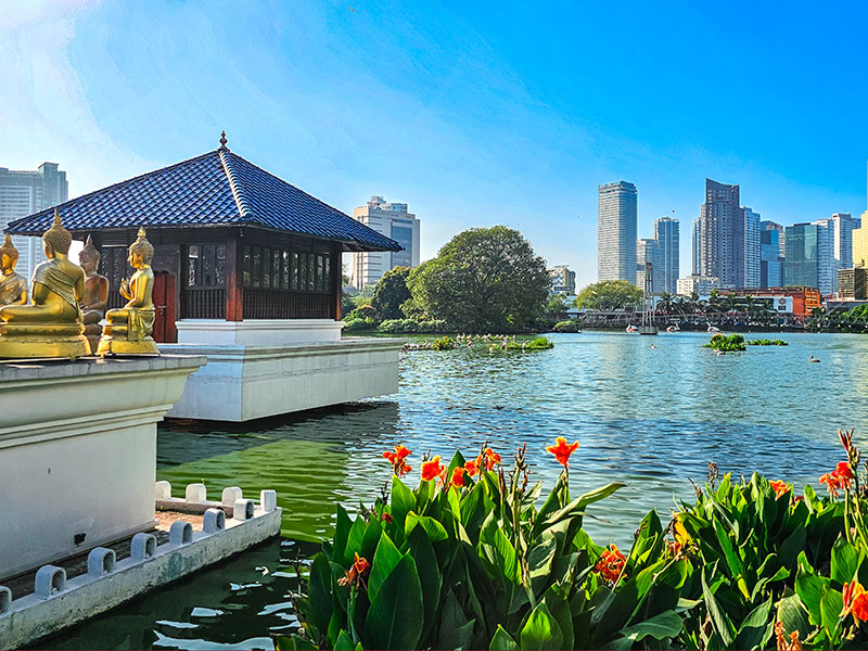 Lakeside pavilion with golden statues in the foreground, orange flowers by the water, and a modern city skyline under a clear blue sky.